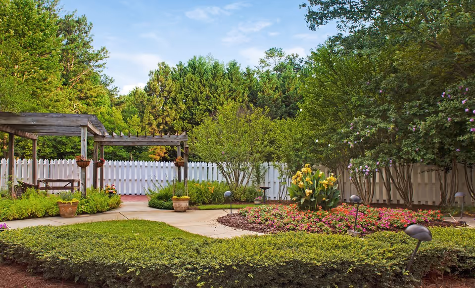 A well-maintained garden area with a variety of green shrubs, flowering plants, and trees. There is a paved walkway winding through the garden, a white picket fence in the background, and a wooden pergola with hanging flower pots on the left side.