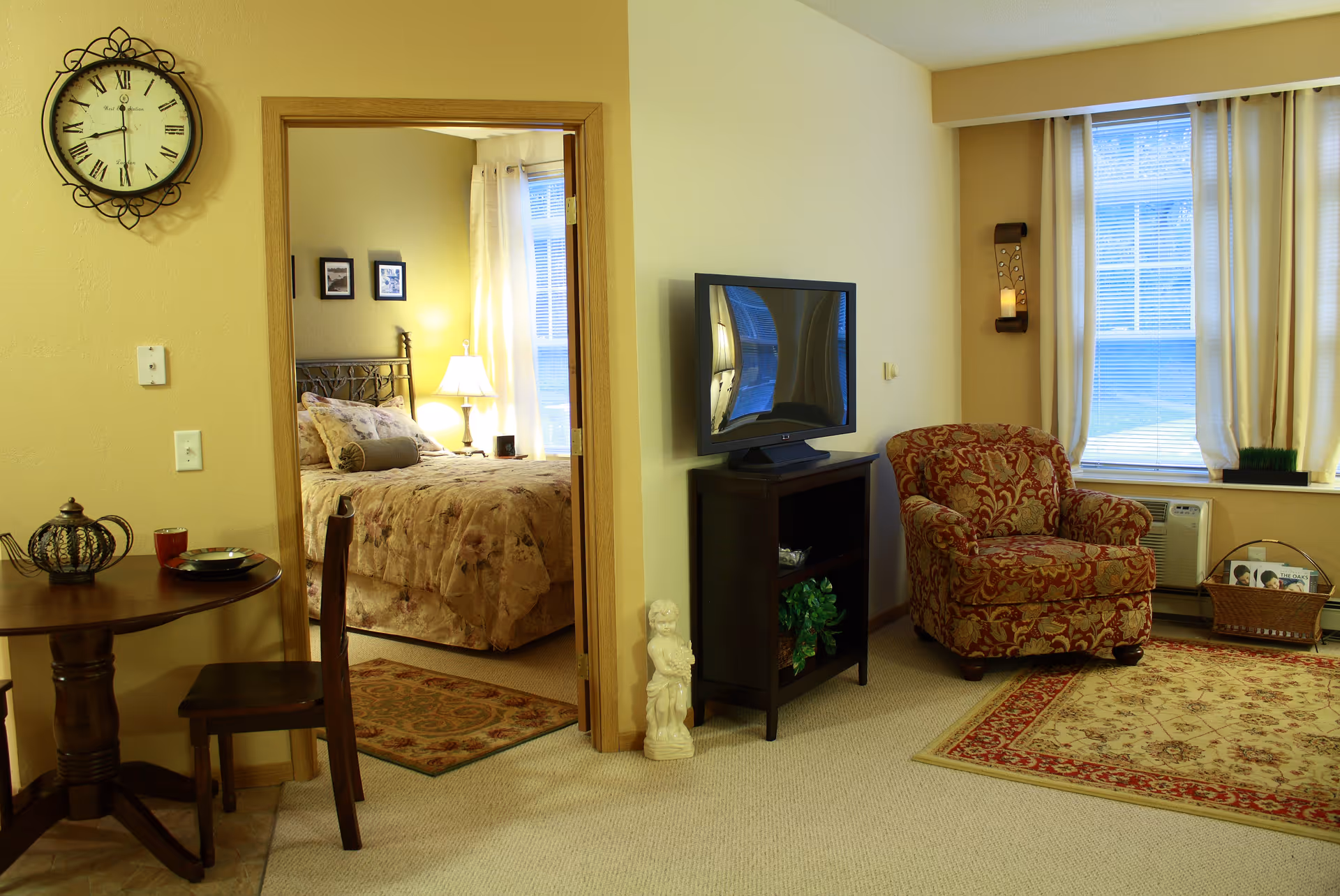 A cozy living area with a patterned armchair, a small TV on a stand, and a window with curtains. Adjacent to the living area is a bedroom with a bed covered in floral bedding, a bedside lamp, and framed pictures on the wall. A small round dining table with a chair, teapot, cup, and plate is also visible. The walls are painted light yellow, and a decorative clock hangs above the dining table.