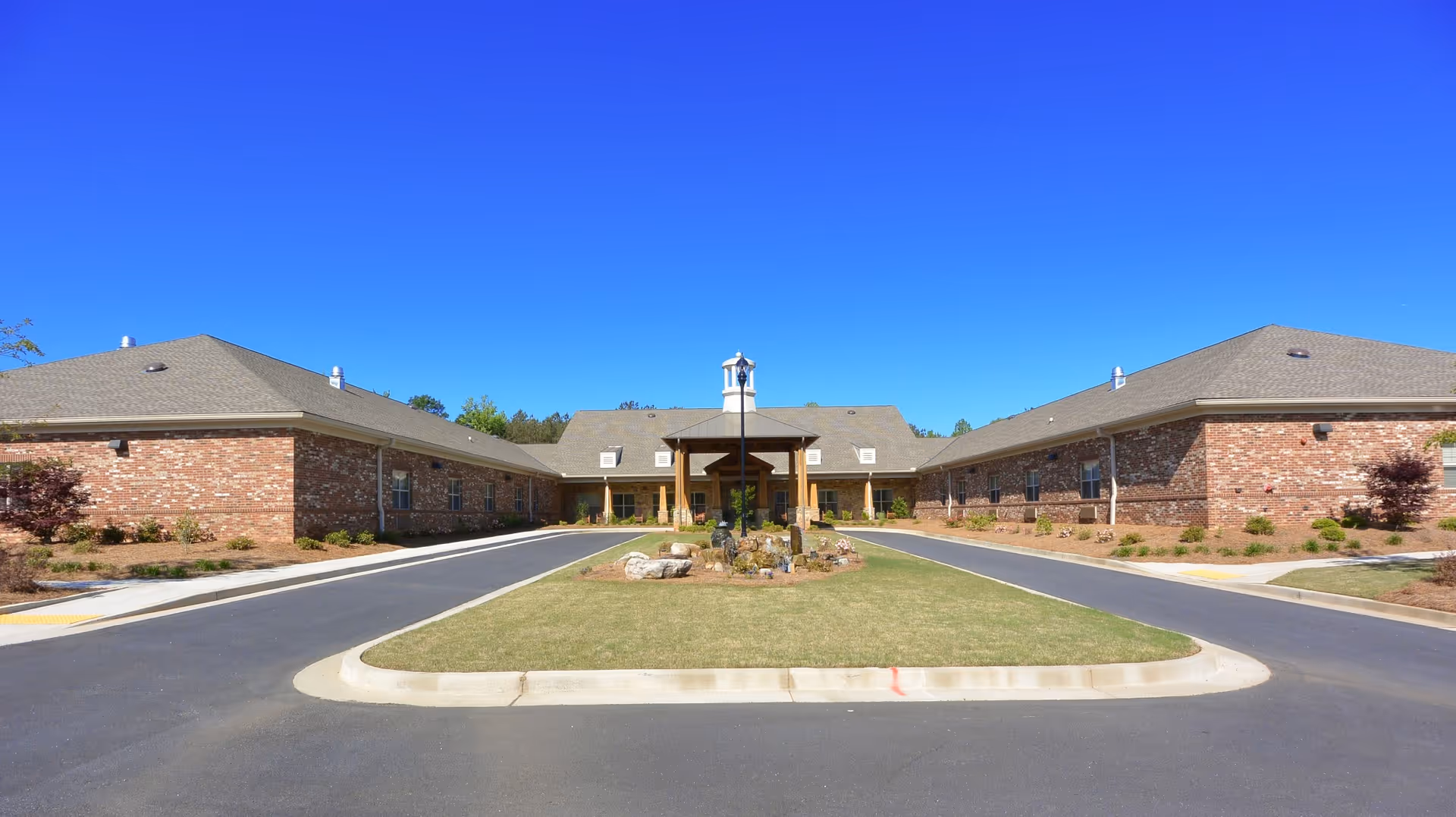 Front exterior view of a single-story brick building with a central covered entrance and a small landscaped island with a bell structure in the driveway, under a clear blue sky.