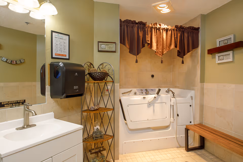 A bathroom with a white sink and faucet on the left, a black paper towel dispenser mounted on the wall, a decorative metal shelf with towels and a basket, and a walk-in bathtub with a brown curtain above it. There is a wooden bench on the right side and beige tiled walls and floor.