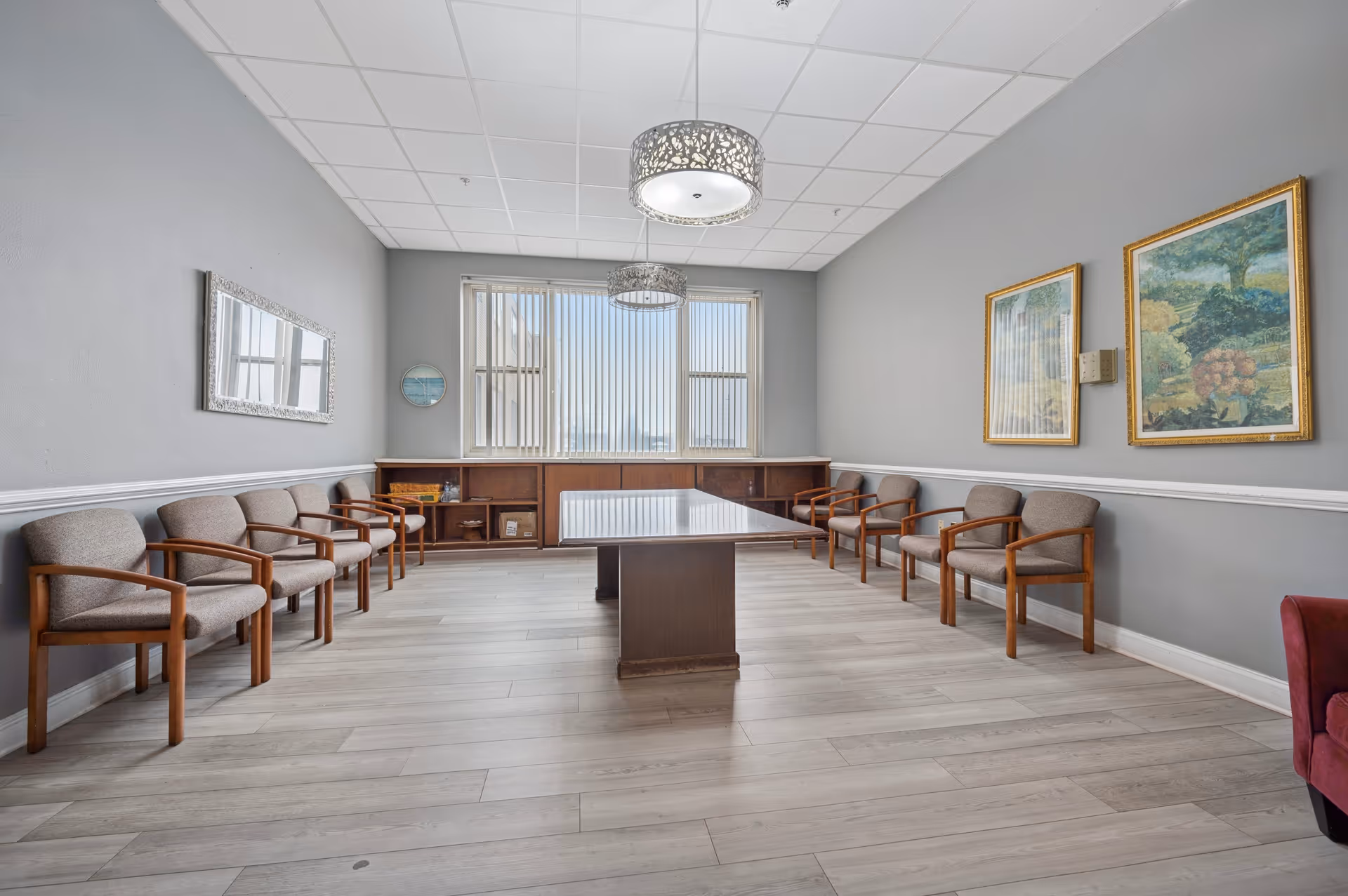 A rectangular meeting or common room with gray walls and light wood flooring. There is a long wooden table in the center, surrounded by cushioned chairs with wooden arms arranged along the walls. Two framed paintings hang on the right wall, and a large mirror is on the left wall. Two decorative ceiling lights hang above the table, and a window with vertical blinds is at the far end of the room.
