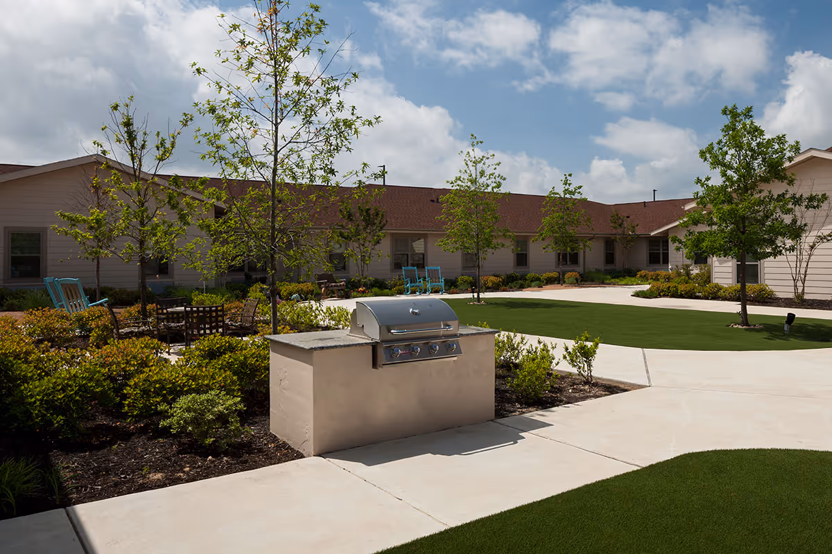Courtyard featuring a built-in outdoor grill, walkways, green lawn, trees, and seating in front of a single-story senior living building.