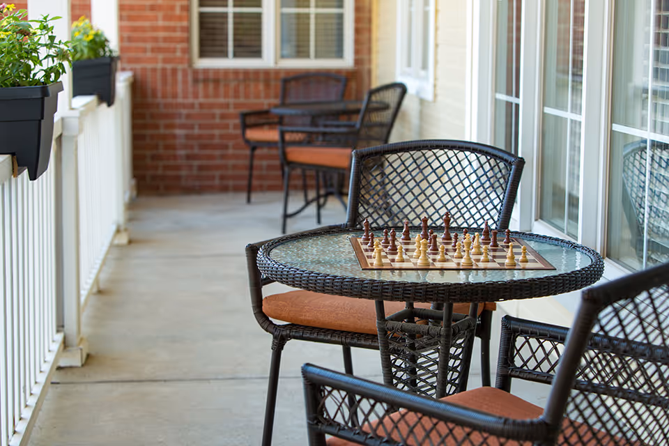 Outdoor patio area with a round glass-top wicker table and wicker chairs with orange cushions. A chessboard with chess pieces is set up on the table. The patio has a white railing with flower boxes containing green plants and yellow flowers. The background shows a brick wall and windows of the building.