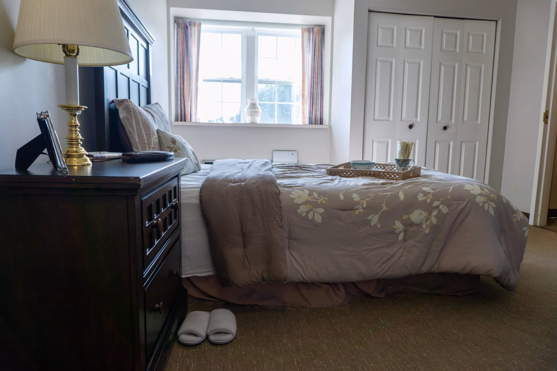 A cozy bedroom with a neatly made bed featuring a floral comforter and pillows. A wooden nightstand with a brass lamp, picture frame, and a book is beside the bed. White slippers are placed on the carpeted floor next to the nightstand. A window with curtains lets in natural light, and there are white double closet doors in the background.