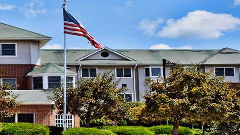 Exterior view of a senior living facility with flagpole, trees, and manicured lawn.