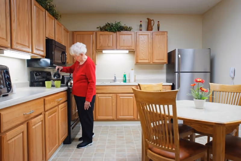 An elderly woman with white hair wearing a red sweater and black pants is pouring coffee into a green cup in a kitchen with wooden cabinets, a stainless steel refrigerator, and a dining table with wooden chairs and a small flower pot with red flowers.