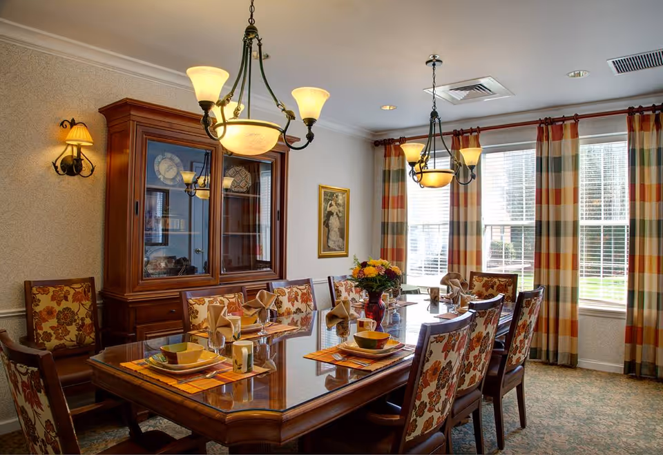 Formal dining room with a long wooden table set for eight, floral-upholstered chairs, a china cabinet and large windows with striped curtains.