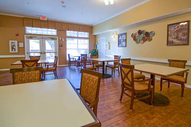 A well-lit dining room with multiple tables and chairs arranged neatly on a wooden floor. The room has large windows and a glass door letting in natural light. The walls are decorated with framed pictures and a decorative plate arrangement.
