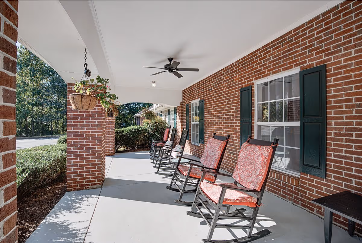 Covered outdoor porch area with several rocking chairs featuring red patterned cushions, hanging plants, brick walls with windows and green shutters, and a ceiling fan overhead.