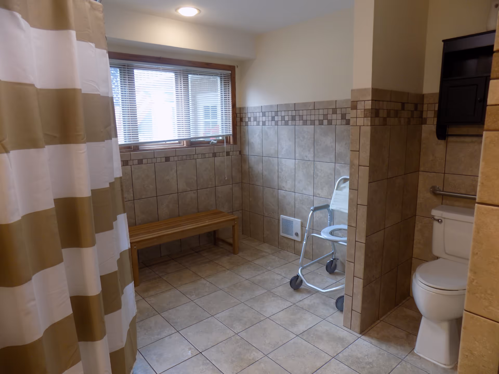 A spacious bathroom with beige and brown tiled walls and floor. There is a white toilet with a grab bar on the right side, a shower area with a beige and white striped curtain on the left, a wooden bench under a window with blinds, and a white shower chair with wheels near the toilet.