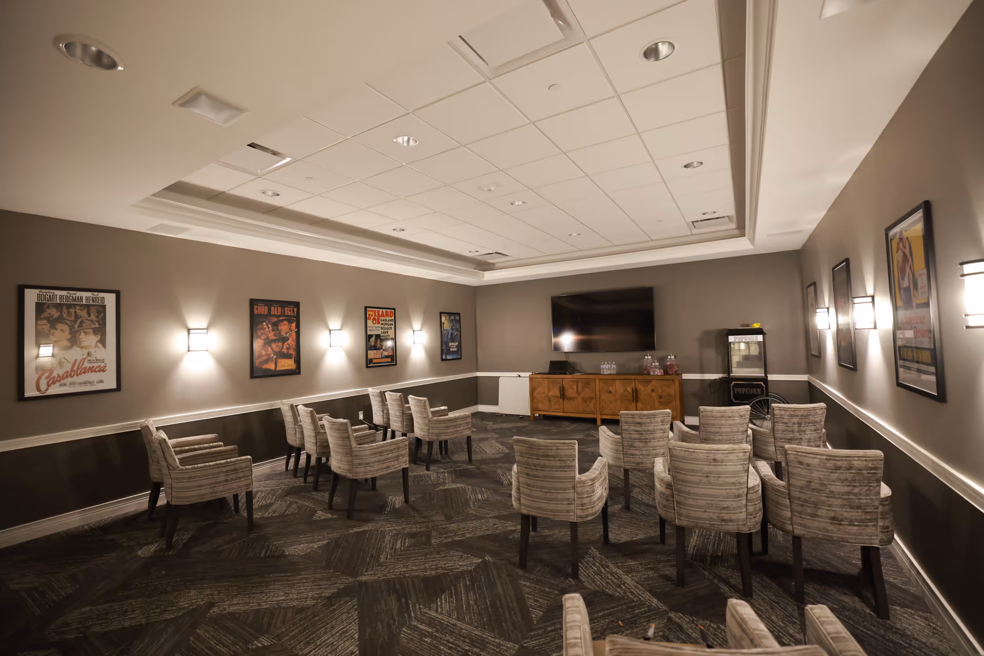 A small theater room with several rows of cushioned chairs facing a large flat-screen TV mounted on the wall. The room has gray walls adorned with framed vintage movie posters and wall sconces providing soft lighting. A wooden cabinet and a popcorn machine are positioned beneath the TV.