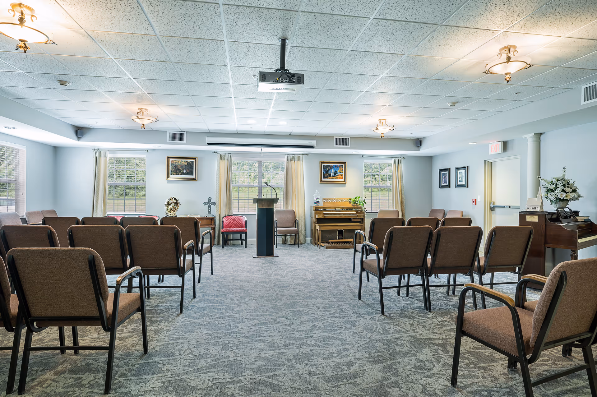 A well-lit room with rows of brown chairs facing a podium and a piano at the front. The room has large windows with beige curtains, framed pictures on the walls, and a carpeted floor. Ceiling lights and a projector are visible overhead.