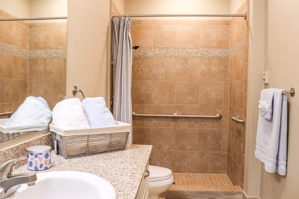 A bathroom with a granite countertop holding a basket of folded towels and a decorative cup. The bathroom features a tiled walk-in shower with grab bars and a shower curtain. A towel rack with a folded towel is mounted on the wall next to the shower.