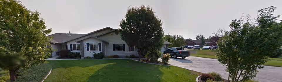 Front exterior of a single-story assisted living building with a manicured lawn, trees, and a driveway with parked vehicles.