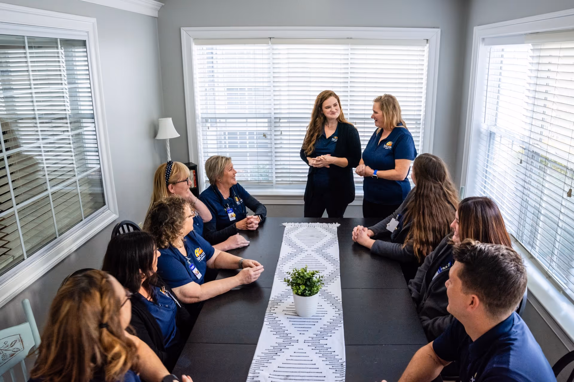 A group of nine people, mostly women, are gathered around a rectangular table in a bright room with large windows and white blinds. Two women are standing at the far end of the table, engaging with the others who are seated. The table has a white patterned runner and a small potted plant in the center. The room has light gray walls and a white lamp in the corner.