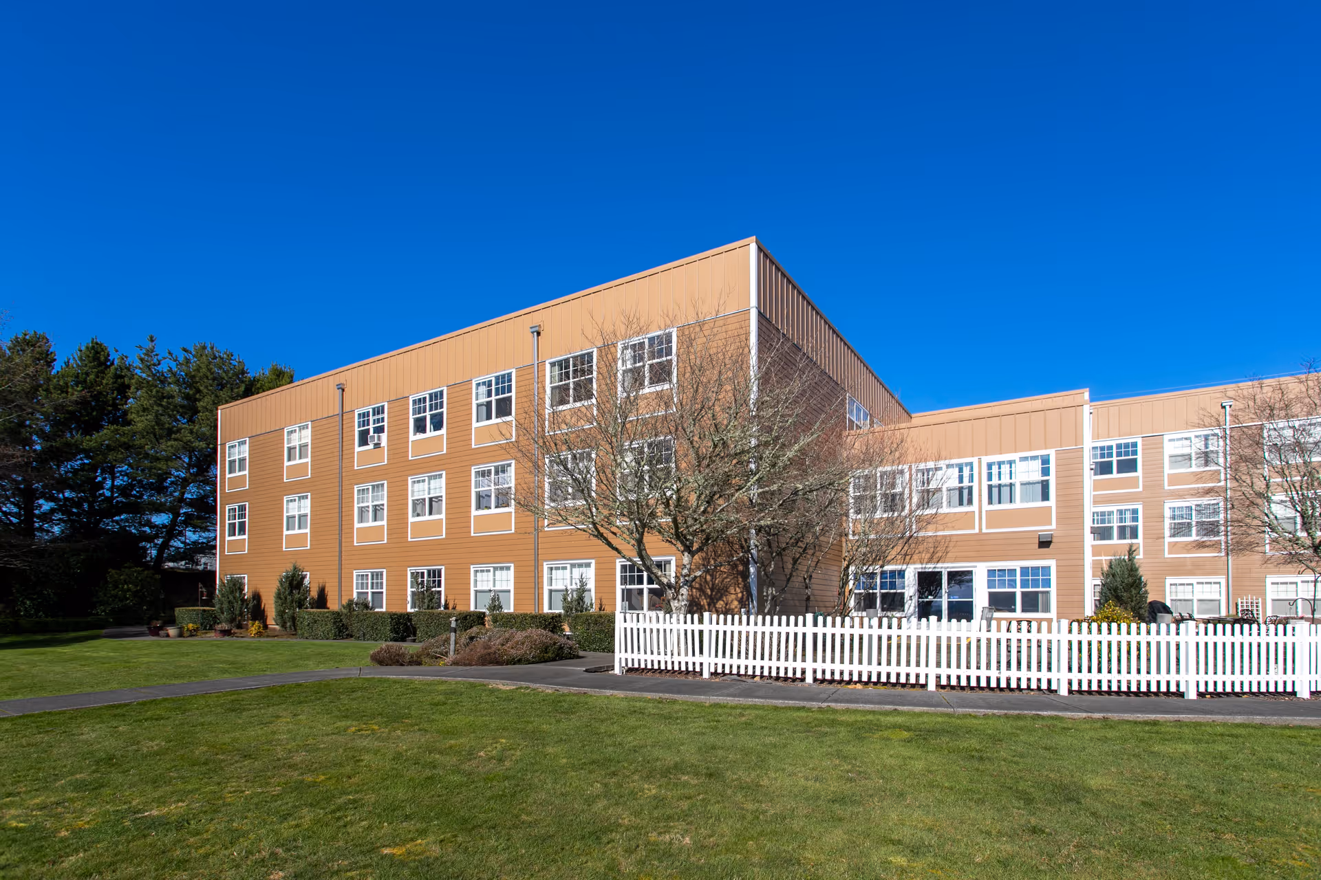 Exterior view of a three-story senior living facility building with tan siding and many windows, surrounded by a well-maintained lawn, a white picket fence, and some trees under a clear blue sky.