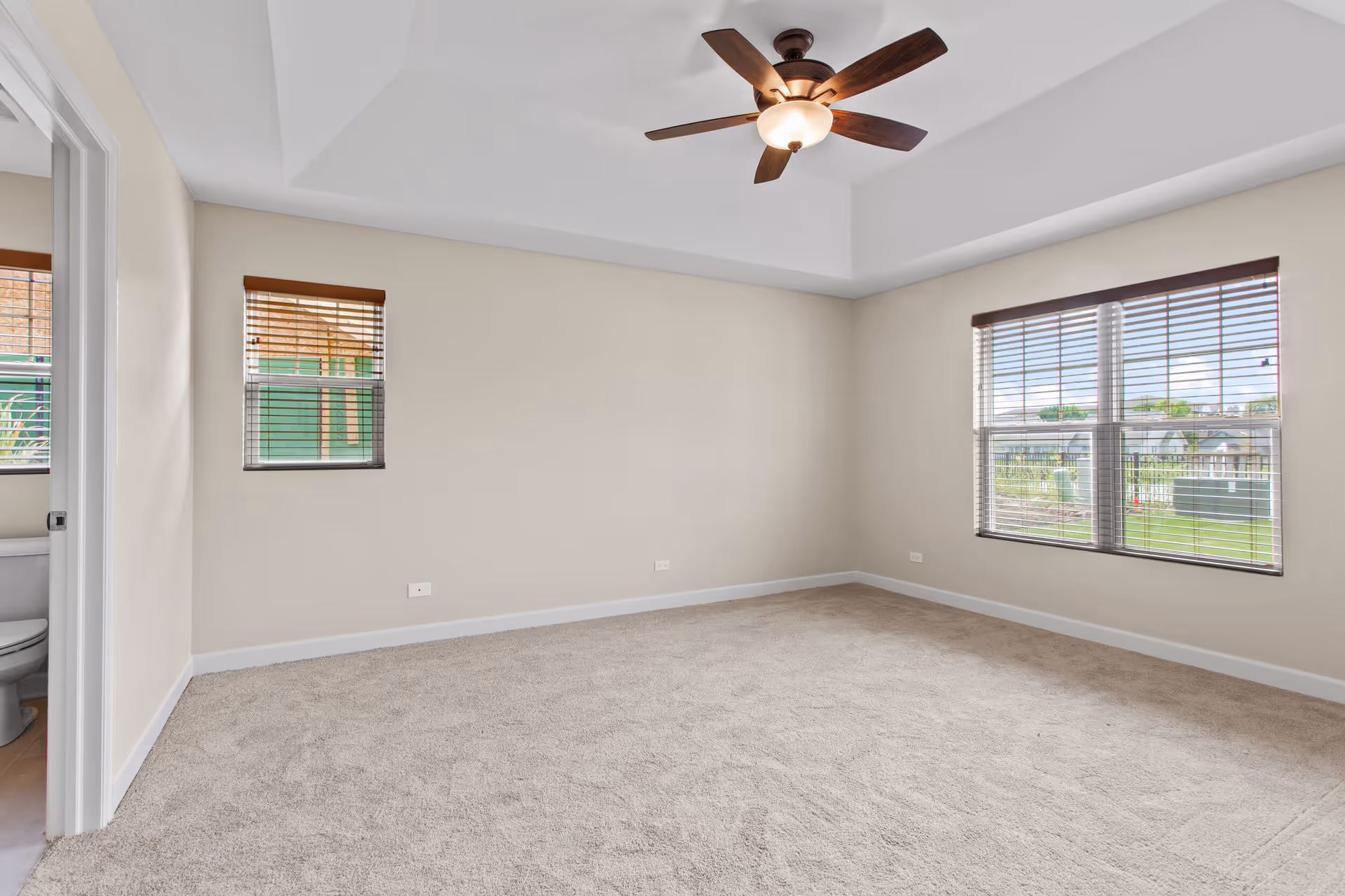 Empty bedroom with beige carpet, light beige walls, two windows with wooden blinds, a ceiling fan with light, and a partial view of an adjacent bathroom.