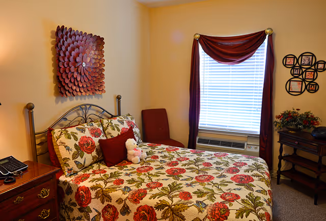 A cozy bedroom with a bed covered in a floral patterned quilt featuring red and green colors. There are matching pillows and a small white teddy bear on the bed. The room has beige walls with a decorative wall hanging above the bed. A window with white blinds and burgundy curtains is on the right side, with an air conditioning unit below it. There is a wooden nightstand with a telephone on the left and a small wooden table with a flower arrangement on the right.