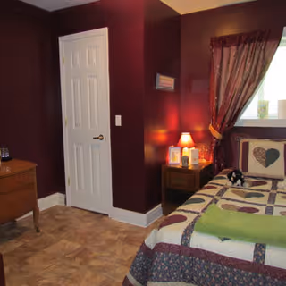 A cozy bedroom with dark red walls, a bed with a quilted heart-patterned bedspread, a green towel folded at the foot of the bed, a small wooden nightstand with a lamp and framed pictures, a window with a curtain, and a closed white door.