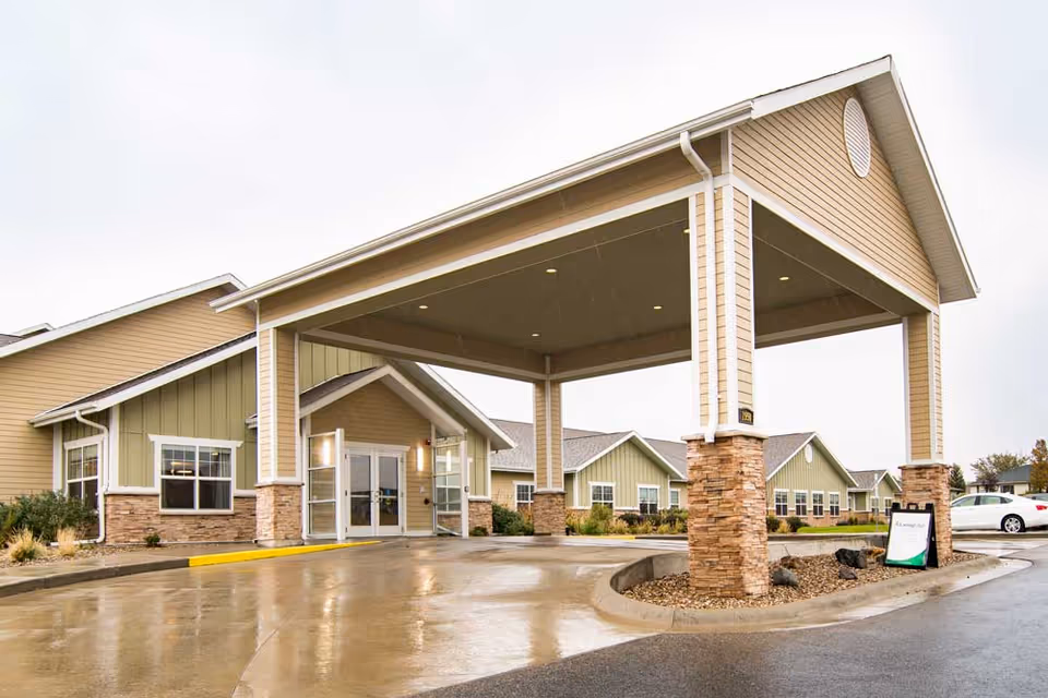 Exterior view of the Edgewood Pierre senior living facility entrance with a covered drop-off area supported by stone pillars. The building has beige and green siding with white trim, and there is a wet driveway in front, indicating recent rain.