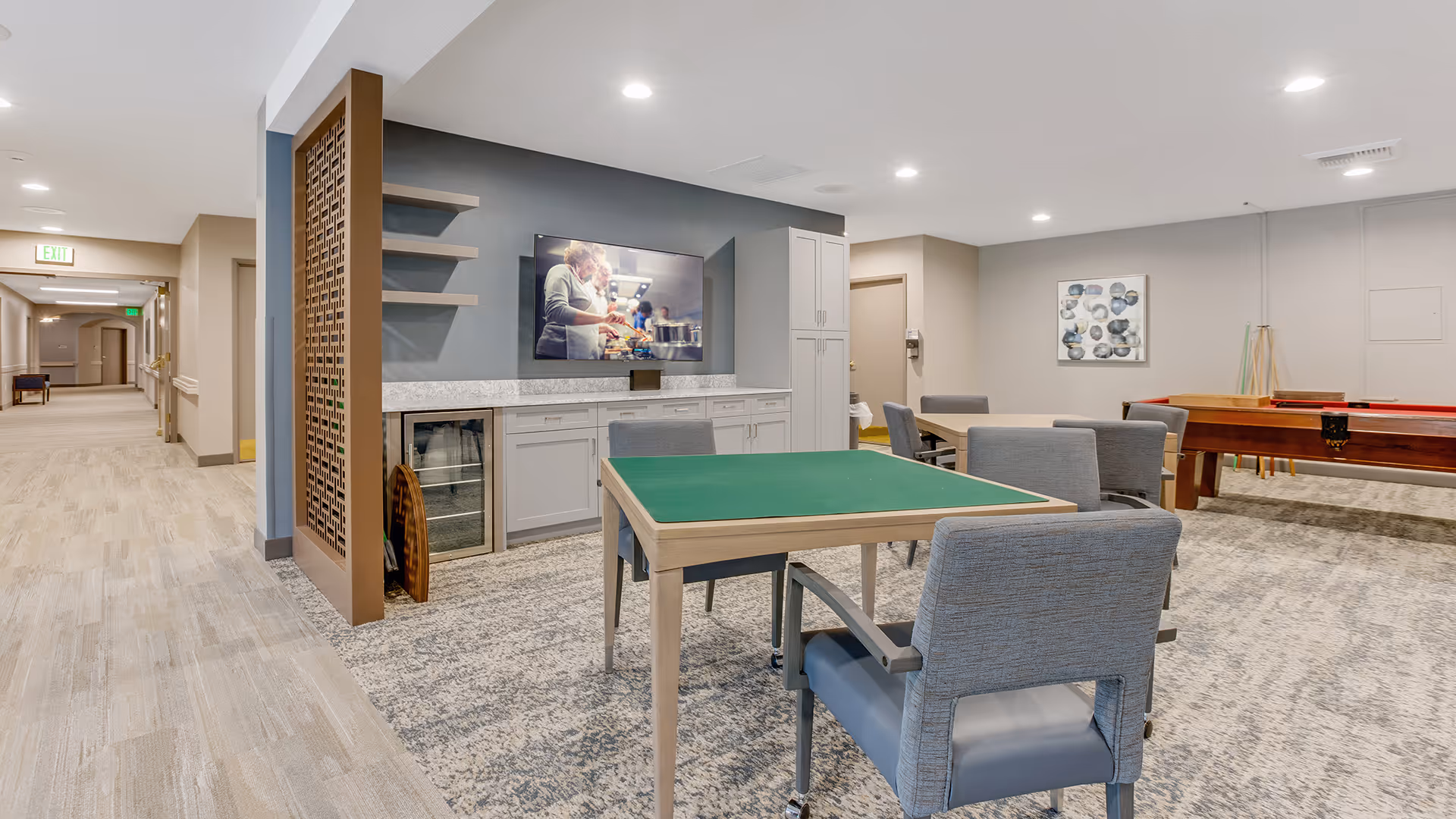 A well-lit recreational room in a senior living facility featuring a green card table with four gray cushioned chairs, a shuffleboard table in the background, a wall-mounted TV displaying a cooking show, and a hallway leading to other rooms.