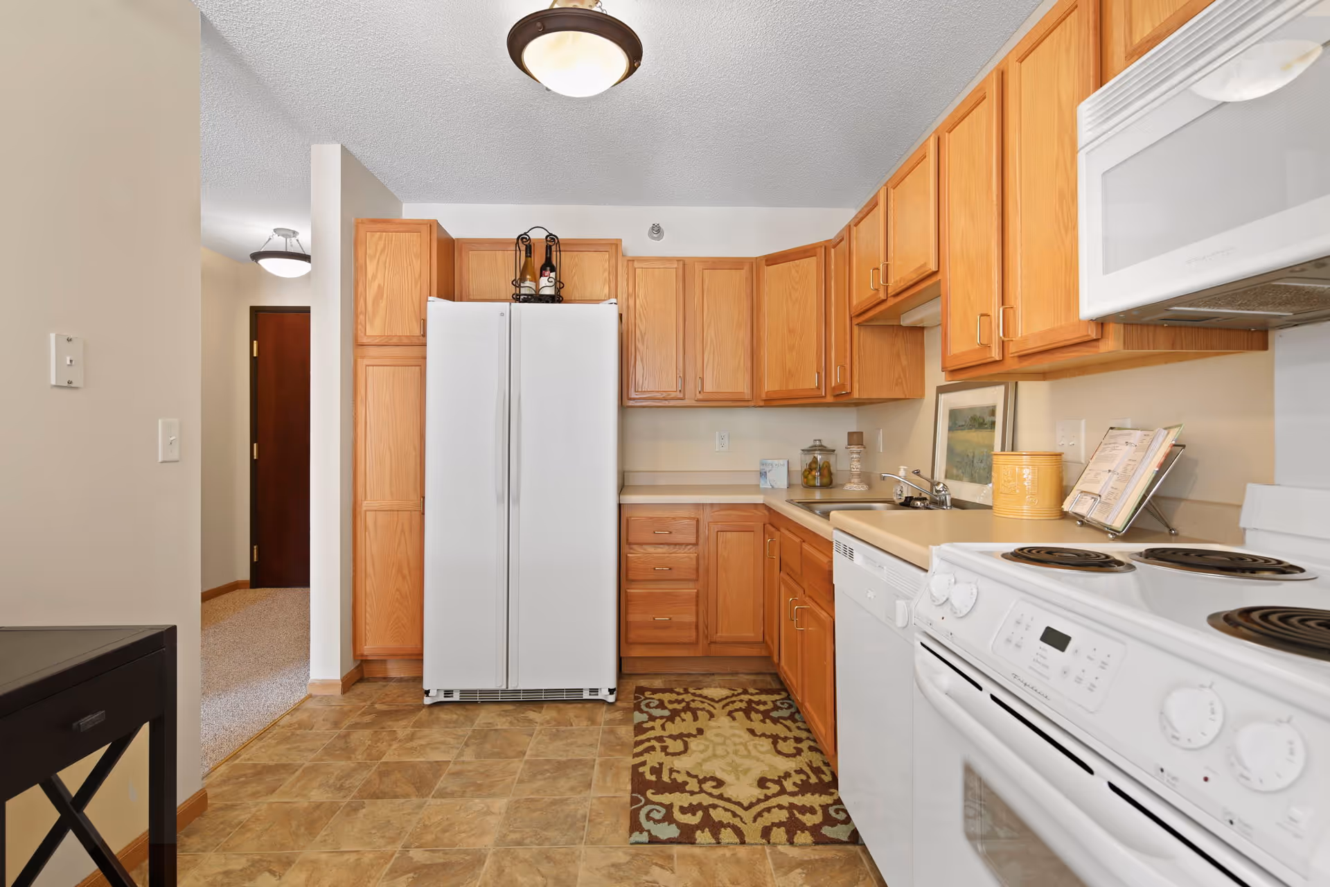 A kitchen with wooden cabinets, a white refrigerator, white electric stove with oven, white microwave, and a white dishwasher. The floor has a patterned rug in front of the sink area. There is a small table visible on the left side and a hallway leading to a door in the background.