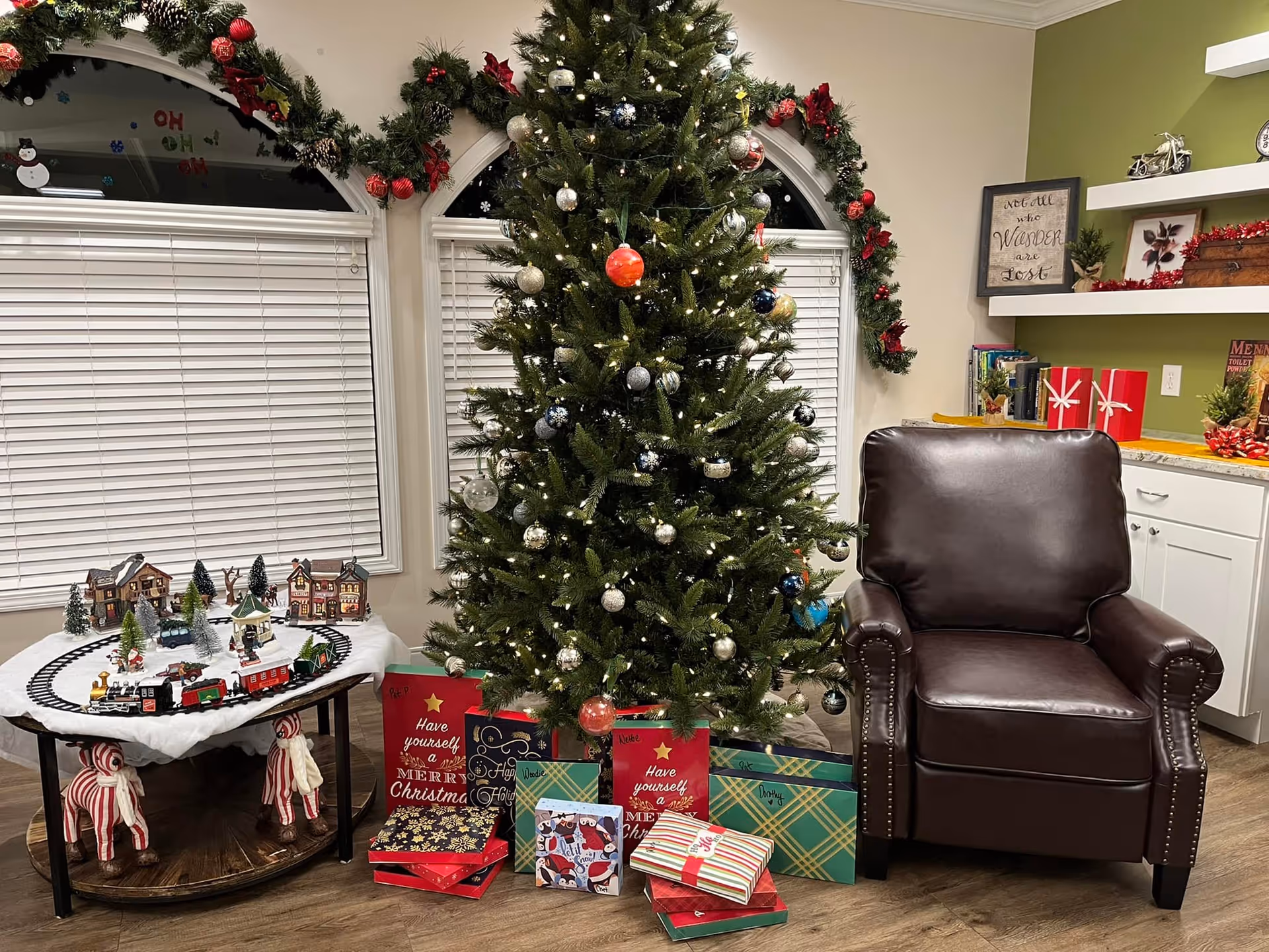 A cozy living room decorated for Christmas with a tall Christmas tree adorned with lights and ornaments. Several wrapped presents are placed under the tree. To the left, there is a table with a miniature Christmas village and a toy train set. The room features two arched windows with white blinds and garlands decorated with red ornaments. To the right, there is a dark brown leather armchair next to white cabinets and shelves with holiday decorations and books.