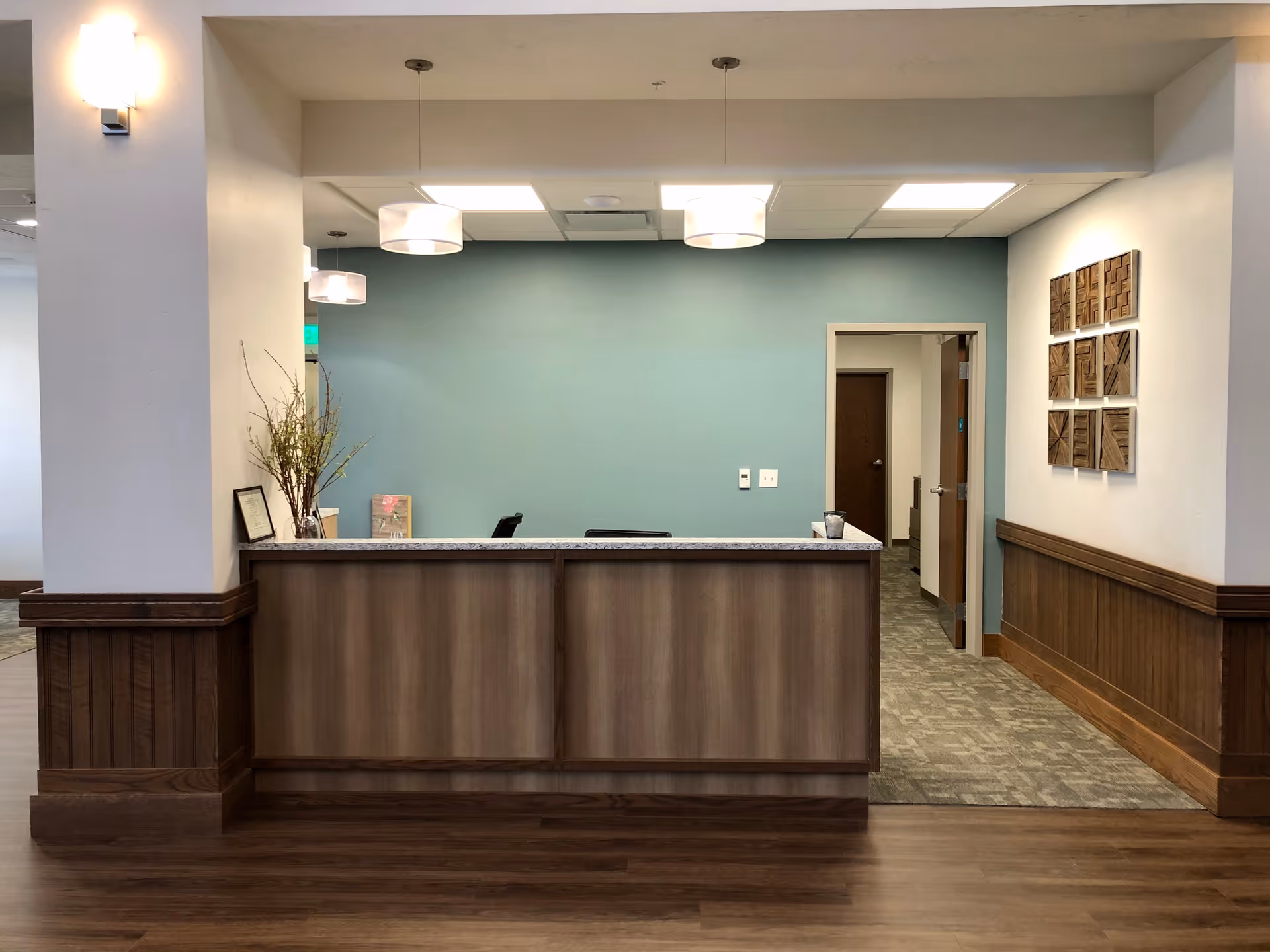 Reception desk area in Meridian Meadows Assisted Living with a wooden counter, a light blue wall behind it, three pendant lights hanging from the ceiling, and a hallway with open doors to the right. There is a decorative plant and framed items on the counter, and wooden wall art on the right wall.