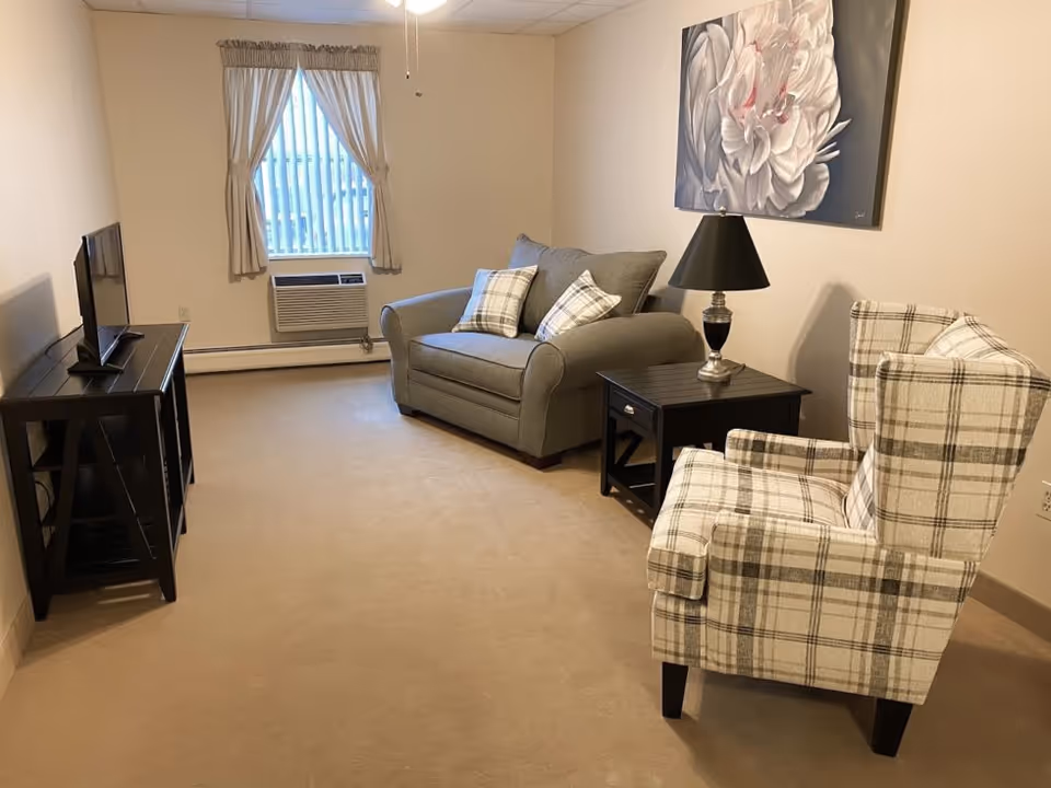 A small living room with a gray loveseat, plaid wingback chair, TV on a stand, side table with lamp, window with curtains, and floral wall art.