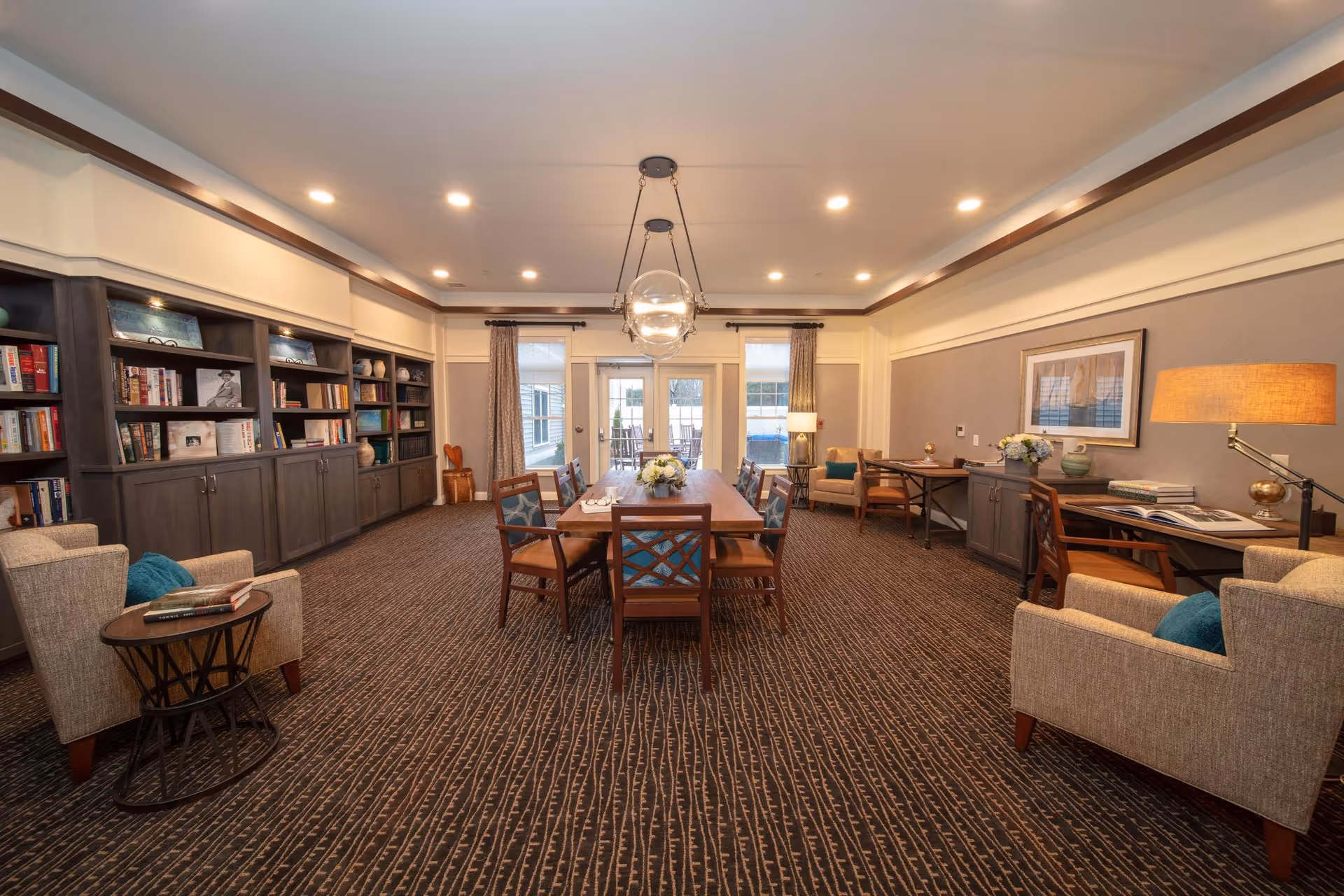 Spacious senior living common room with a central dining table, bookshelves, armchairs, and large windows at the far end.