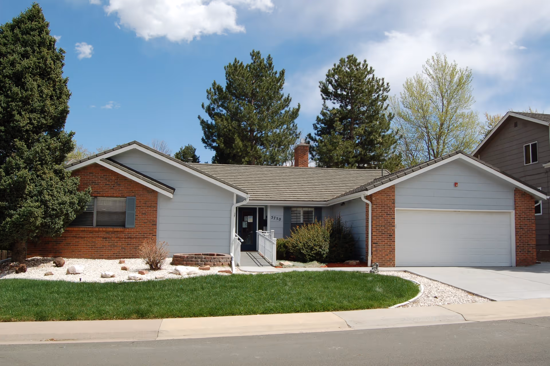 Single-story residential building with a brick and light gray exterior, a two-car garage, a small ramp leading to the front door, green lawn, and trees in the background under a partly cloudy sky.