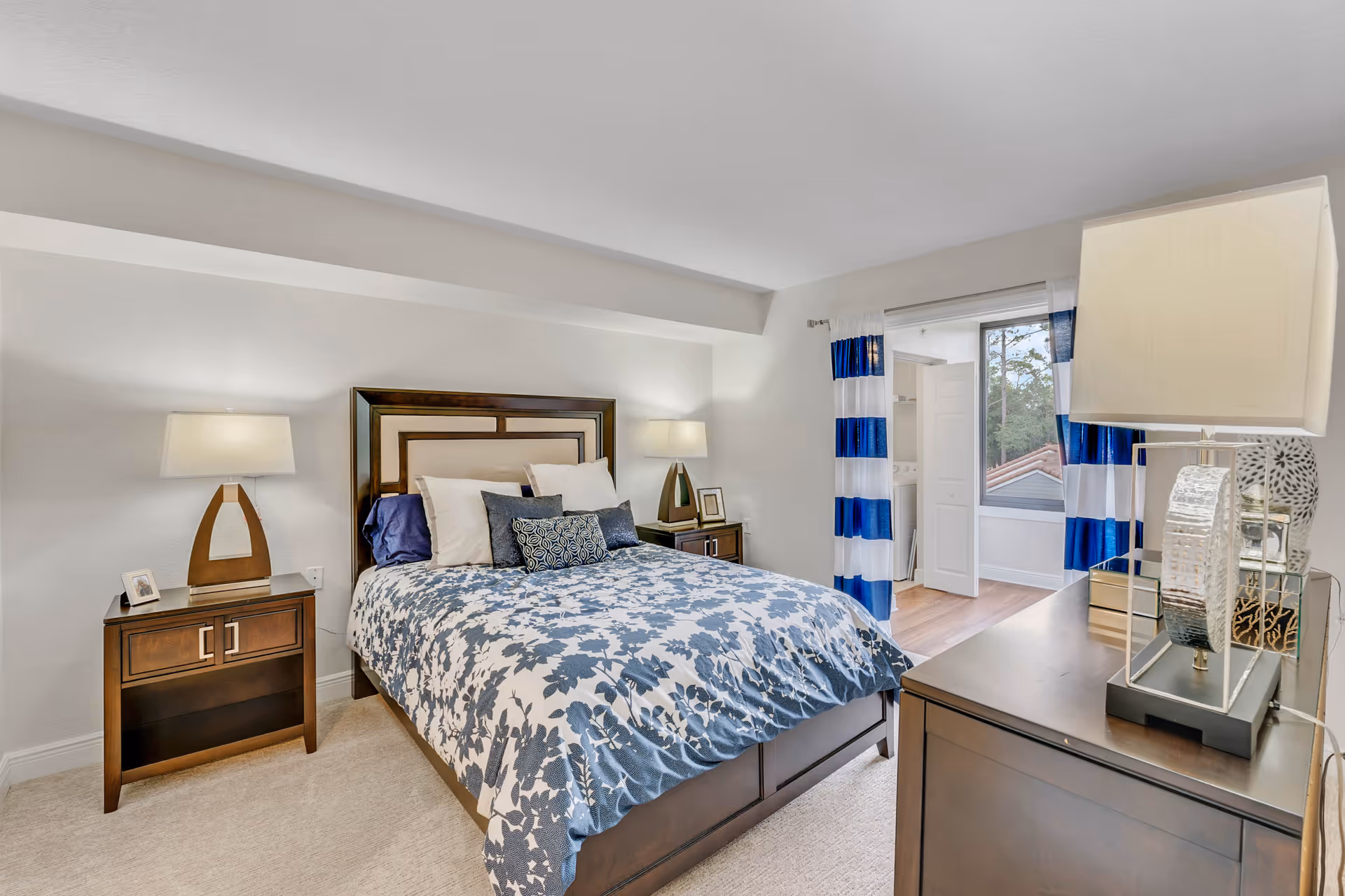 Bright bedroom with a queen bed dressed in blue-and-white floral bedding, two nightstands with lamps, a dresser, and a window with blue-striped curtains.