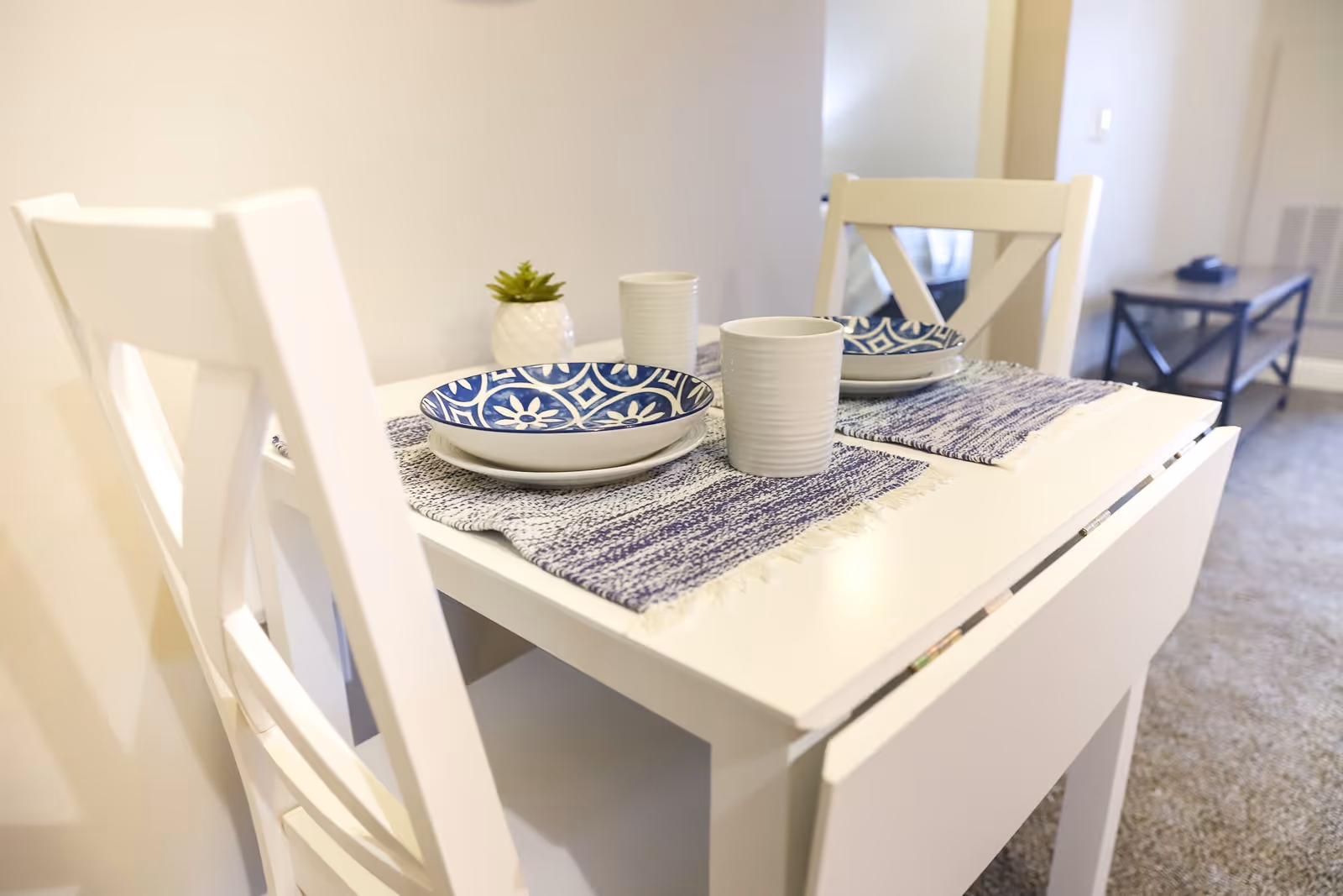 A small white dining table set for two with blue and white patterned plates, white cups, and blue woven placemats. Two white chairs are positioned at the table, and a small green succulent plant in a white pot is placed on the table. In the background, there is a glimpse of a living area with a coffee table and carpeted floor.