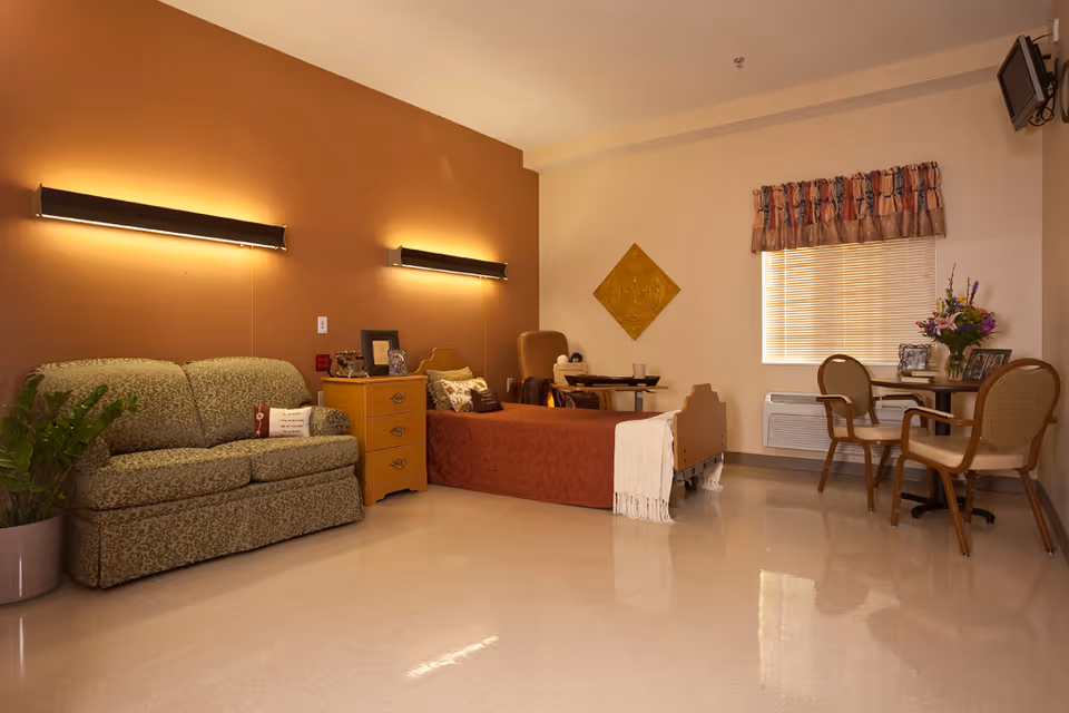 A cozy room with a single bed covered in a rust-colored blanket, a green patterned loveseat, a wooden nightstand with framed photos, a small dining table with two chairs, a window with blinds and a valance, and a wall-mounted TV in the corner.