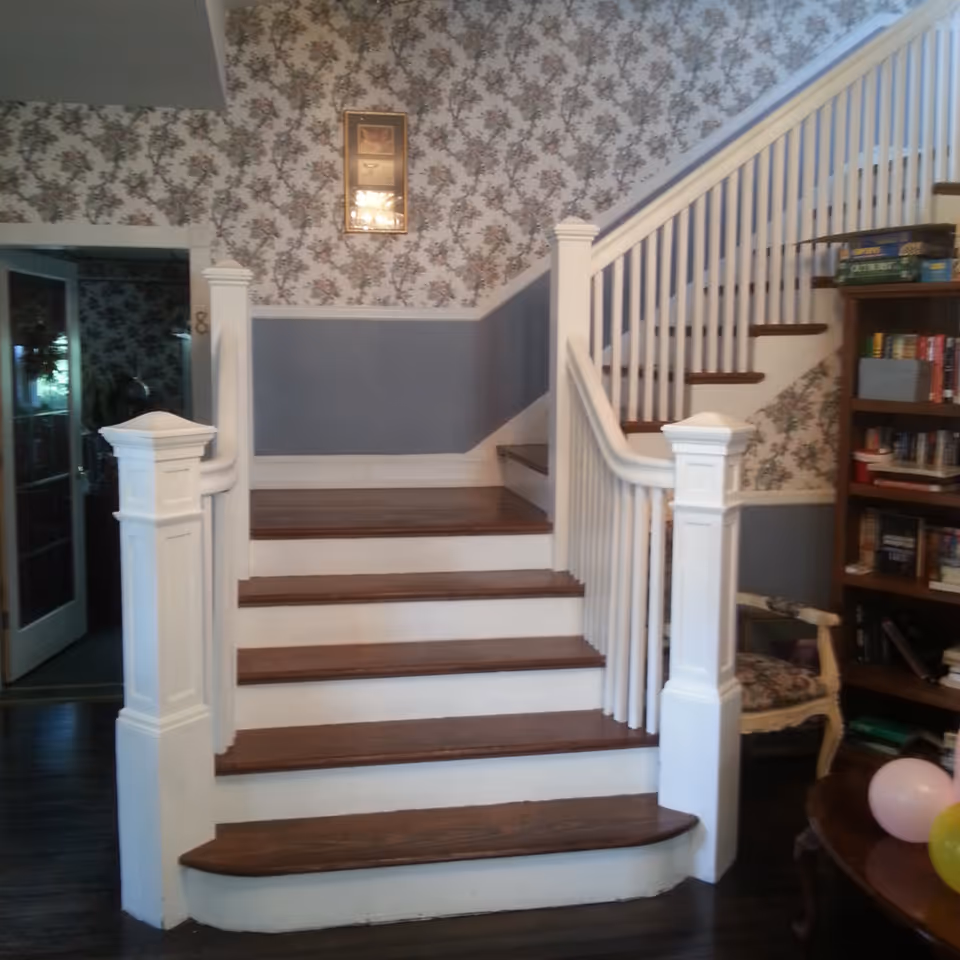 Interior view of a staircase with wooden steps and white railings in a home-like setting. The walls are decorated with floral wallpaper and a framed picture hangs above the stairs. To the right, there is a bookshelf filled with books and board games, and a cushioned chair is partially visible. A small table with balloons is also seen in the lower right corner.
