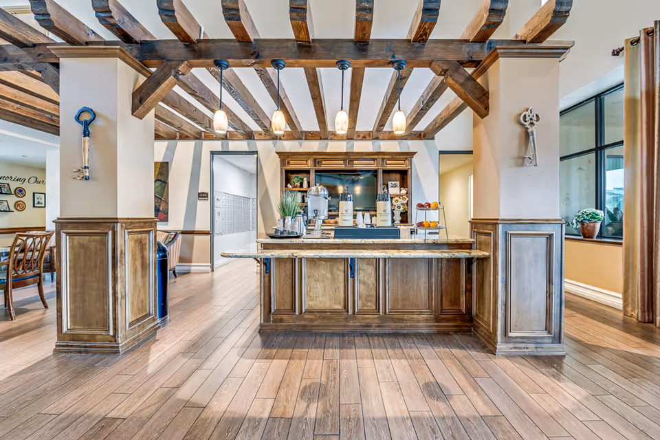 Interior view of a senior living facility common area with wooden beams on the ceiling, a wooden counter with beverages and snacks, pendant lights hanging above, and decorative keys mounted on the pillars. There are chairs and tables visible in the background along with large windows and curtains.