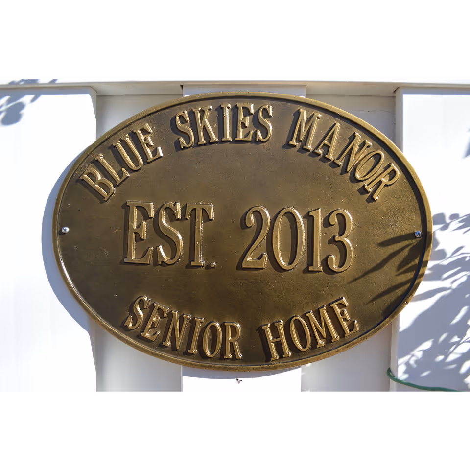 Bronze oval plaque reading "Blue Skies Manor EST. 2013 Senior Home" mounted on a white exterior surface.