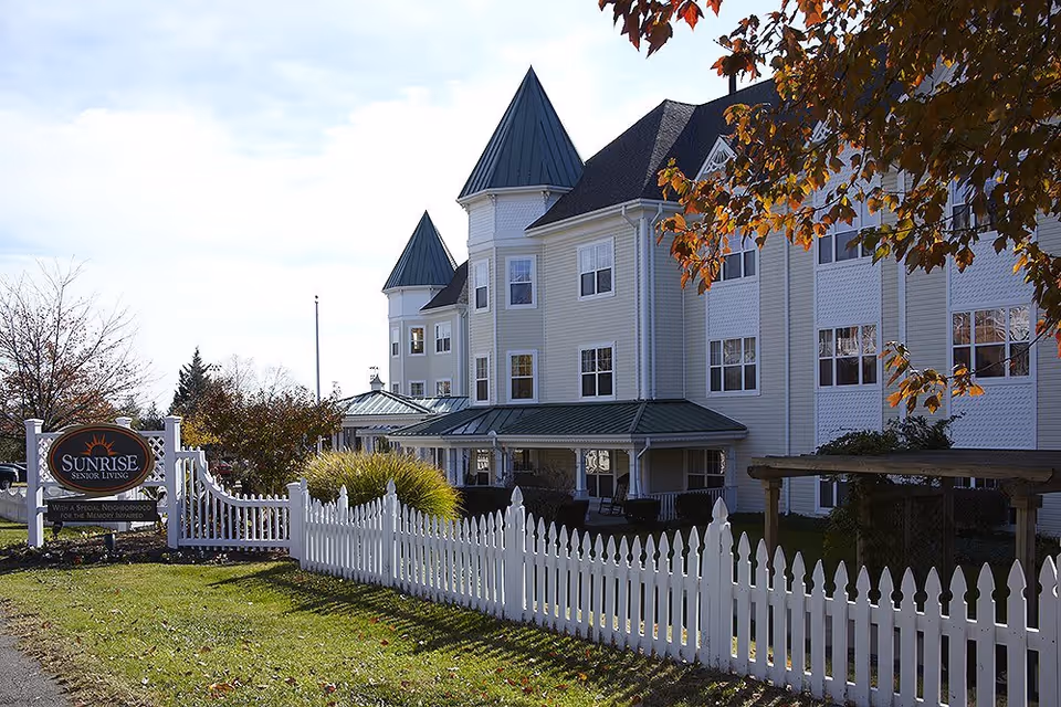 Exterior view of Sunrise Senior Living facility with a white picket fence, green lawn, and a large building featuring multiple windows and pointed roof towers. There are trees with autumn-colored leaves in the foreground.