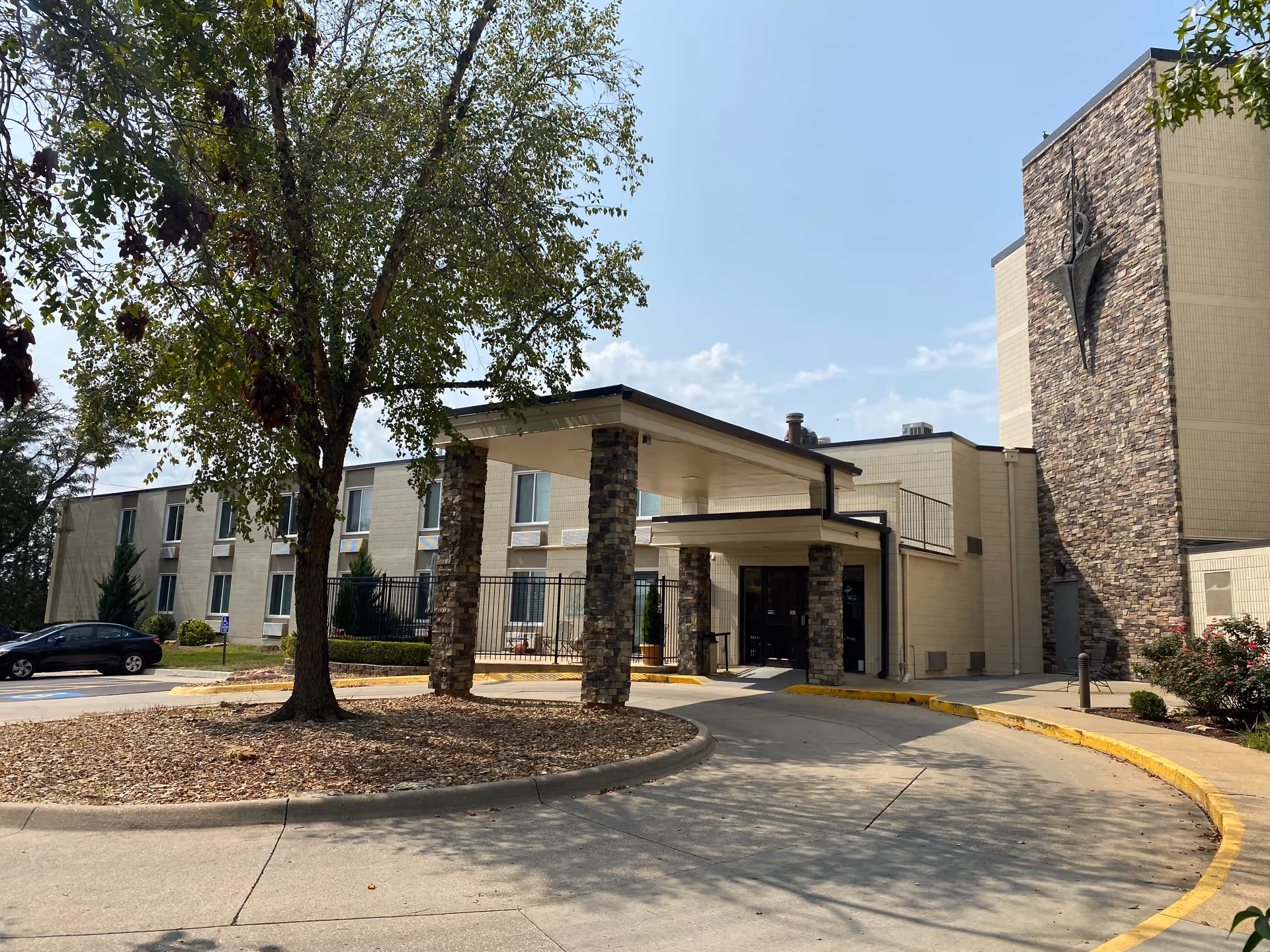 Exterior view of Lawrence Presbyterian Manor showing the main entrance with a covered drop-off area supported by stone pillars. The building is two stories tall with beige walls and a stone accent wall featuring a decorative metal sculpture. There is a tree and some landscaping in front of the entrance, and a car is parked to the left.