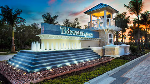 Illuminated entrance sign for Tidewater by Del Webb at dusk, featuring a tiered water fountain, palm trees, and a small tower structure with a peaked roof, set against a colorful sky.