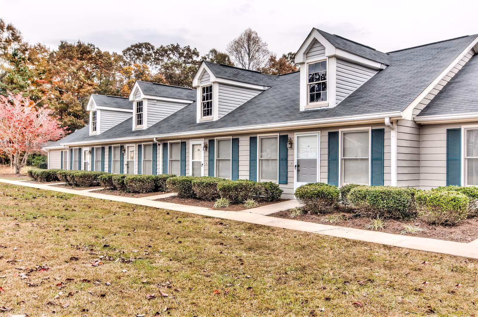 Exterior view of a single-story residential building with multiple units, each having white doors and windows with blue shutters. The building has a gray roof with dormer windows and is surrounded by neatly trimmed bushes and a grassy lawn with a concrete walkway. Trees with autumn foliage are visible in the background.
