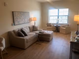 A cozy living room with a beige sectional sofa, two patterned armchairs near a window, a small round table between the chairs, two table lamps on either side of the room, a large decorative wall art above the sofa, and wooden flooring.