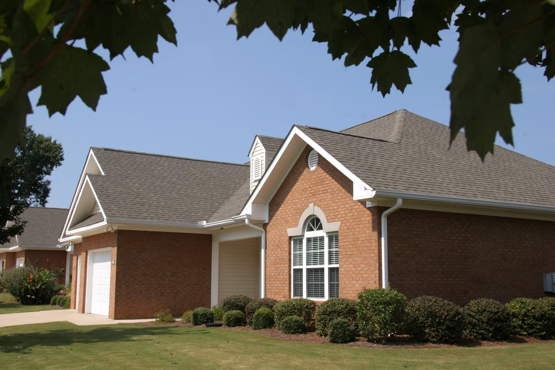 Exterior view of a single-story brick building with a gable roof, white trim, and a large window with white shutters. The building is surrounded by neatly trimmed bushes and a well-maintained lawn, with tree leaves partially framing the top of the image.