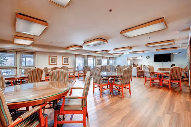 A spacious dining room with multiple round wooden tables and striped upholstered chairs arranged neatly. The room has large windows allowing natural light to enter, wooden flooring, and ceiling light fixtures. A flat-screen TV is mounted on the far wall, and framed artwork is displayed on the walls.