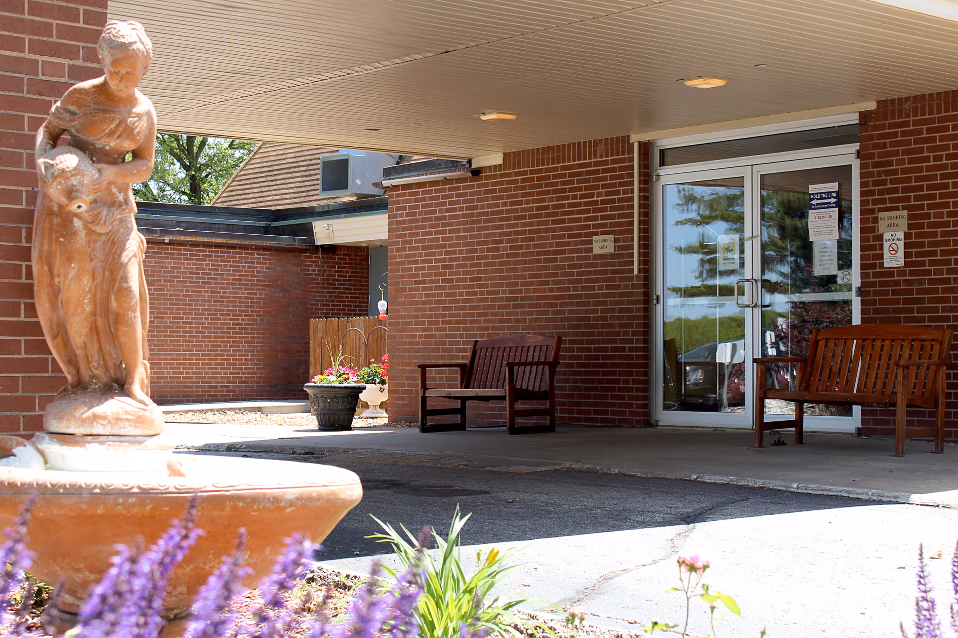 Covered front entrance of a brick nursing home showing a statue fountain, wooden benches, potted flowers, and glass double doors.