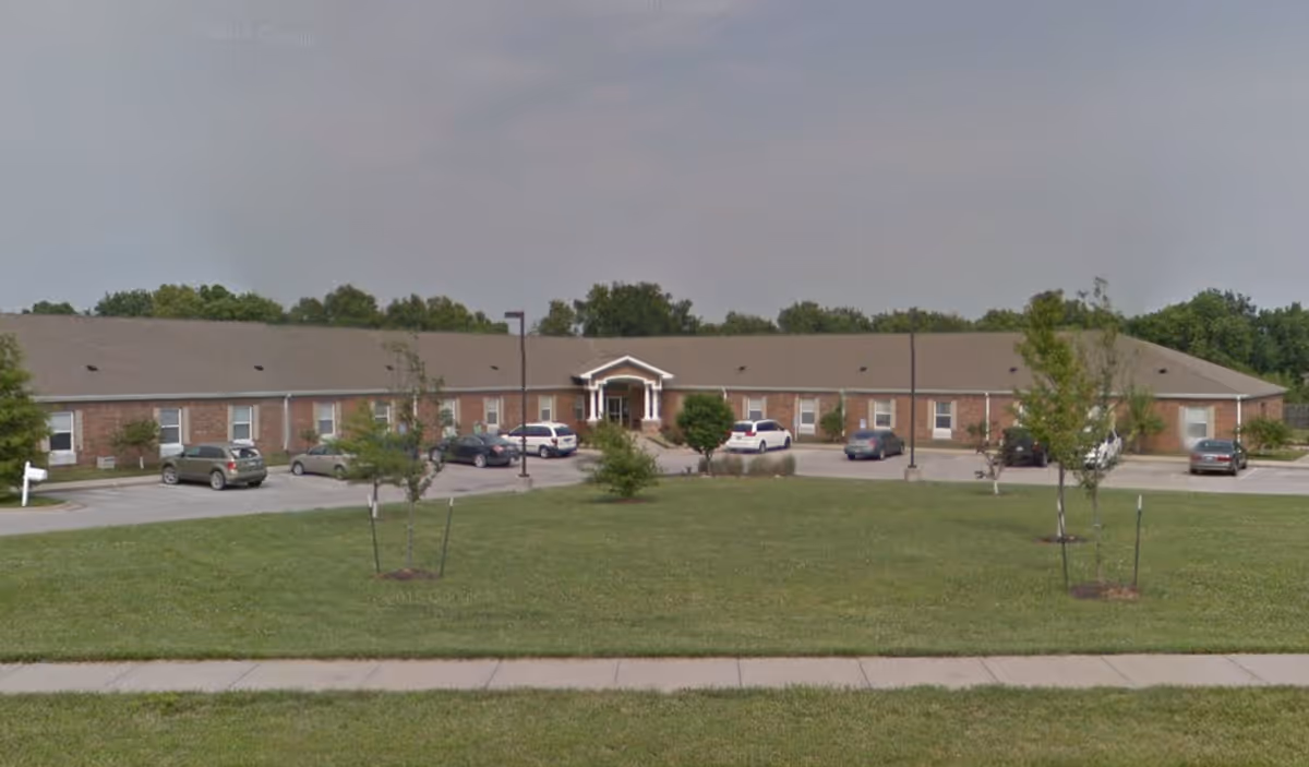 Front exterior view of a single-story brick senior living facility building with a pitched roof, several windows, a central entrance with a small portico, a parking lot with several cars, and a grassy lawn with young trees in front.