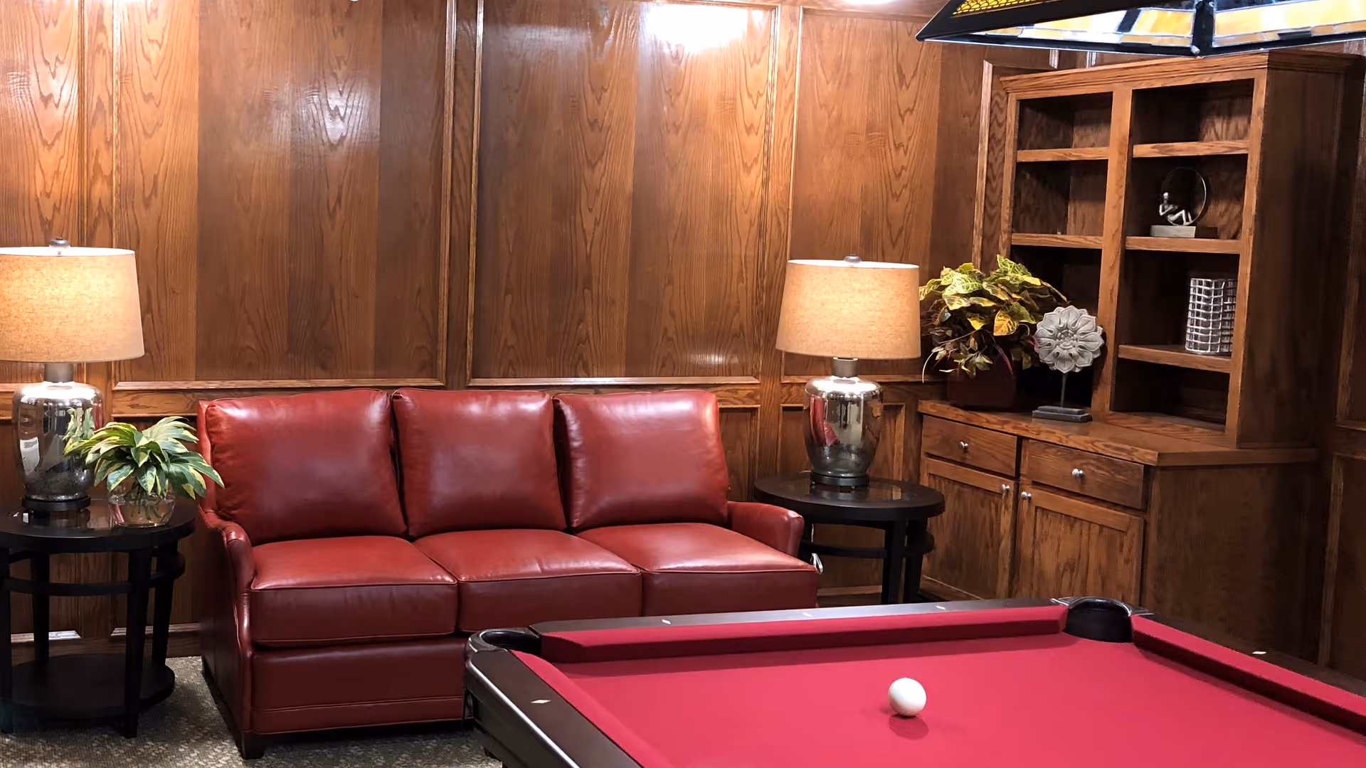 A cozy room with wood-paneled walls featuring a red leather sofa flanked by two side tables with lamps and plants. In front of the sofa is a red pool table with a white cue ball on it. To the right, there is a wooden cabinet with shelves holding decorative items.