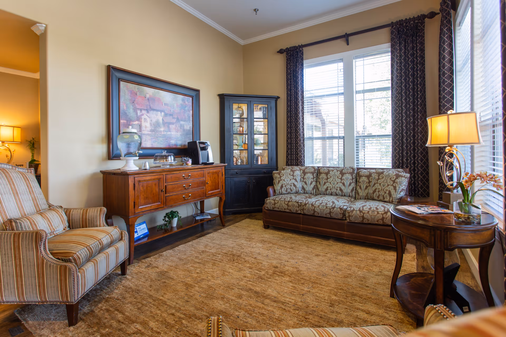 A cozy living room with a patterned sofa, a striped armchair, a wooden sideboard with a water dispenser and coffee maker, a glass-front cabinet, a round wooden side table with a lamp and flowers, large windows with dark patterned curtains, and a beige carpet.