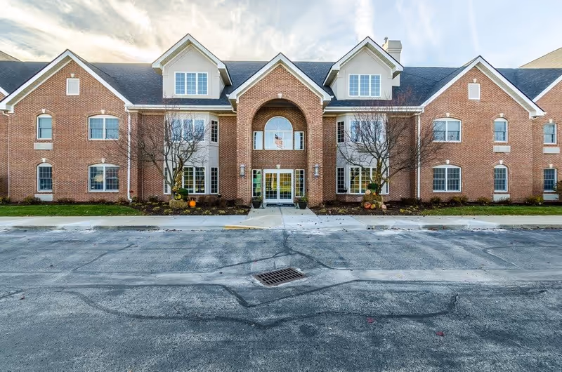 Front exterior view of a large brick assisted living facility building with multiple windows, a central entrance with an arched design, and a paved driveway in front. There are small trees and landscaping near the entrance.