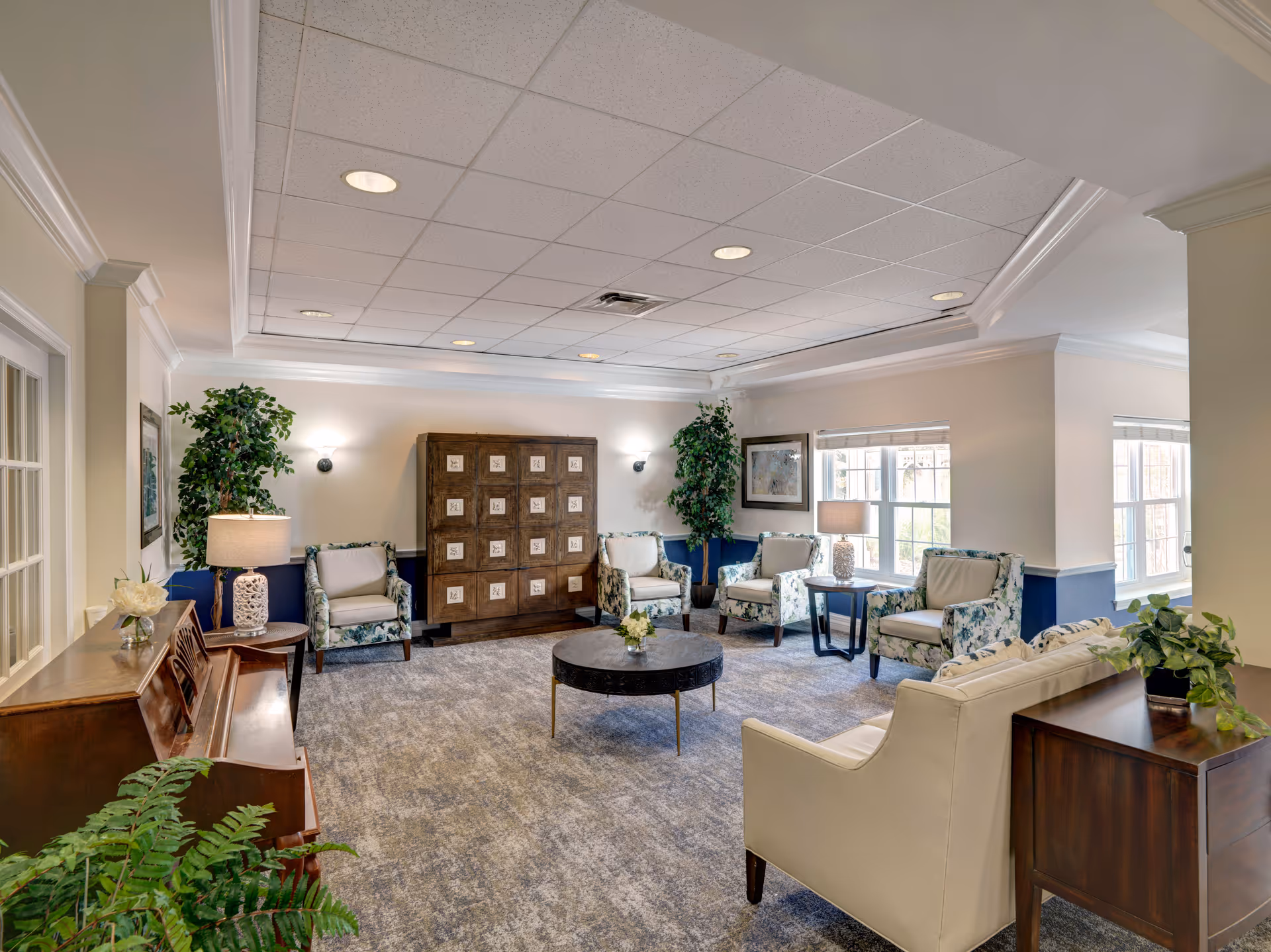 A bright and spacious living room area with multiple upholstered armchairs arranged around a round coffee table. The room features large windows letting in natural light, a wooden piano on the left, potted plants, table lamps, and a decorative wooden cabinet against the far wall. The walls are painted in a light color with a blue accent on the lower half.